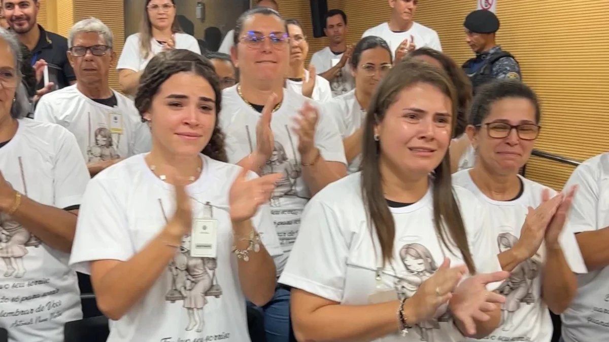 FOTOGRAFIA COLORIDA MOSTRA FAMILIARES APLAUDINDO. ELES ESTÃO VESTIDOS COM BLUSAS COM ARTE QUE HOMENAGEIA A VITIMA. 