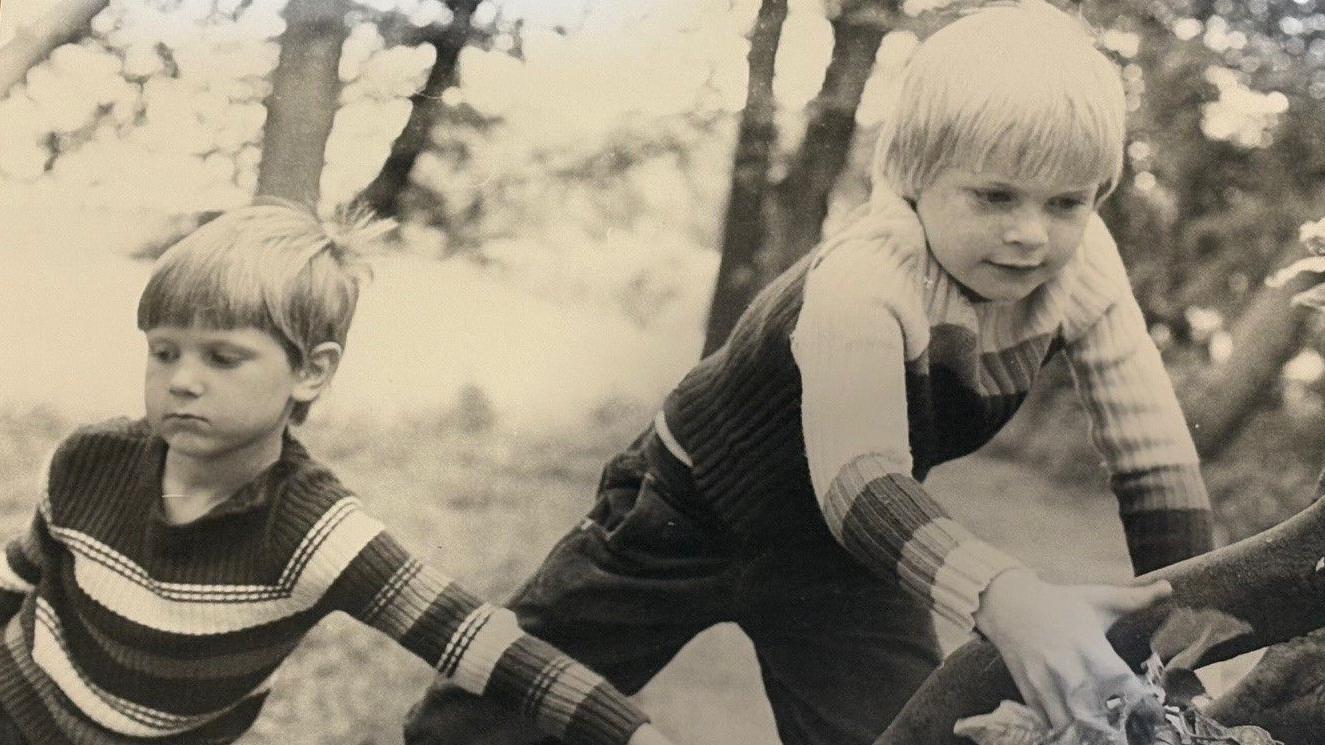 Dois meninos, ambos usando suéteres de listras horizontais, estão ao lado de uma árvore. Um deles a segura, enquanto o outro, com expressão determinada, está prestes a escalá-la