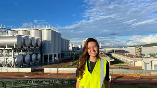 Letícia Lemos Martins veste um colete amarelo refletivo e sorri, com a fábrica da Heineken em Passos, Minas Gerais, ao fundo. Ela tem cabelos ondulados compridos e veste uma camiseta preta por debaixo do colete