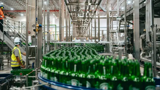 Linha de produção da Heineken em Passos, Minas Gerais, com centenas de garrafas verdes típicas da marca de cerveja. À esquerda, há um operário de colete reflexivo amarelo e capacete branco, com o rosto voltado para o lado oposto ao da câmera