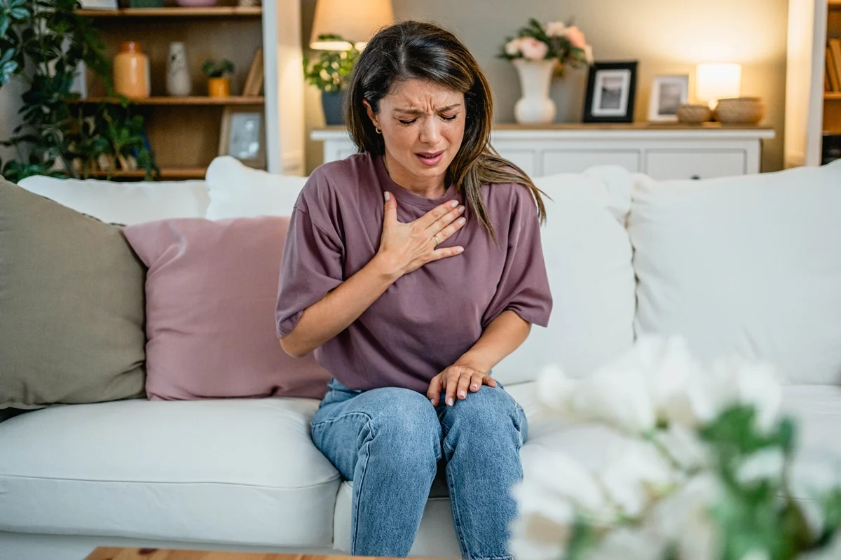 Foto colorida de mulher sentada em sofá e com a mão no peito - Metrópoles