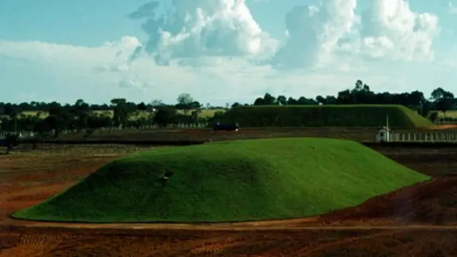 Paisagem aberta com solo avermelhado e vegetação ao redor, destacando dois montes cobertos por grama bem aparada, de formato arredondado, cercados por uma área isolada; ao fundo, há árvores, uma pequena construção e céu com nuvens.